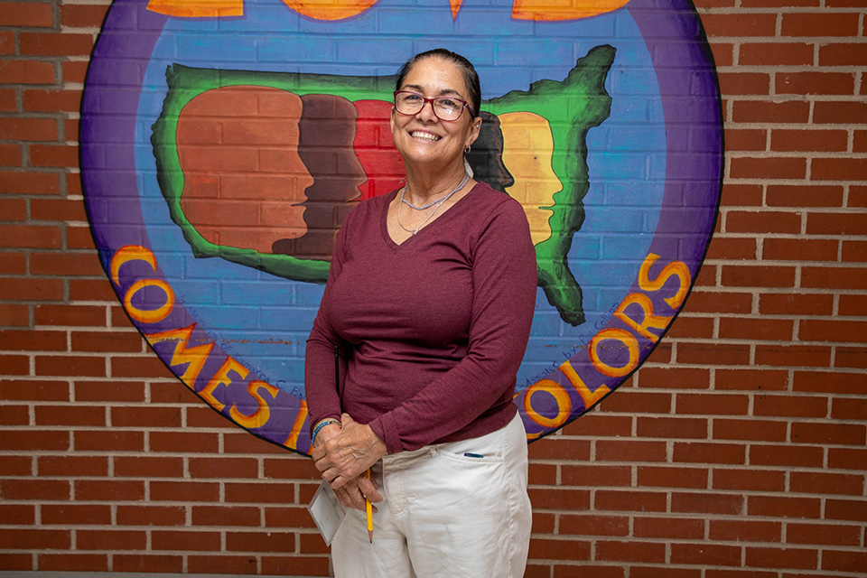 A woman with red glasses, a red shirt and khakis smiles in front of a colorful mural